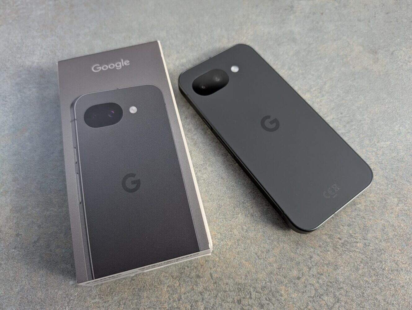 Een Google Pixel smartphone, uit een recente Google Pixel 10a review, ligt naast zijn doos op een grijs oppervlak. De achterkant van de telefoon is zichtbaar en toont de camera en het Google-logo.