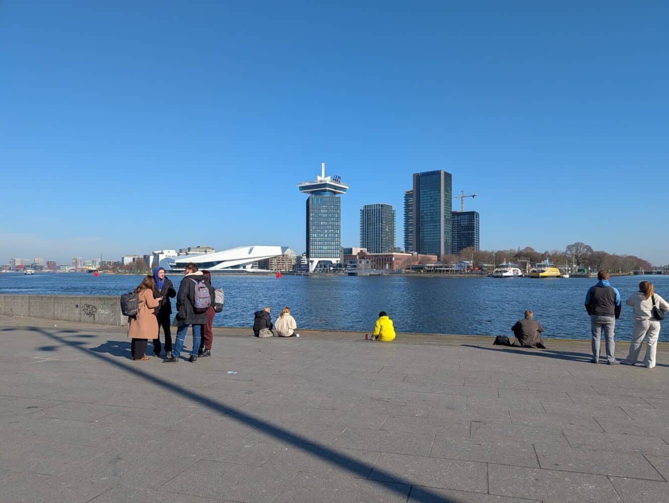 Mensen staan en zitten langs een promenade aan het water met moderne gebouwen en een rivier op de achtergrond onder een strakblauwe hemel, en leggen het levendige tafereel vast - perfect voor foto's die je zou zien in een Google Pixel 10a review.