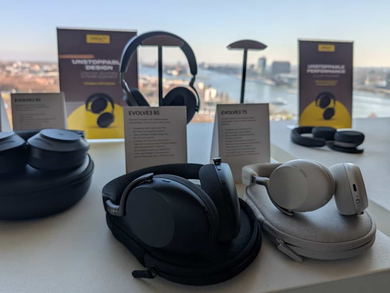 Verschillende modellen Jabra Evolve hoofdtelefoons worden tentoongesteld op een tafel met productinformatiekaarten, met uitzicht op een stadslandschap door een groot raam, vergelijkbaar met opstellingen die te zien zijn in sommige Google Pixel 10a review showcases.