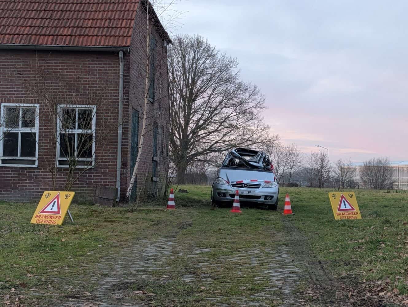 Een zwaar beschadigde auto staat geparkeerd naast een bakstenen gebouw, omringd door verkeerskegels en waarschuwingsborden met de tekst "Brandweer Oefening", die lijken op scènes die te zien zijn in voor- en nafoto's van een Google Pixel 10a review.