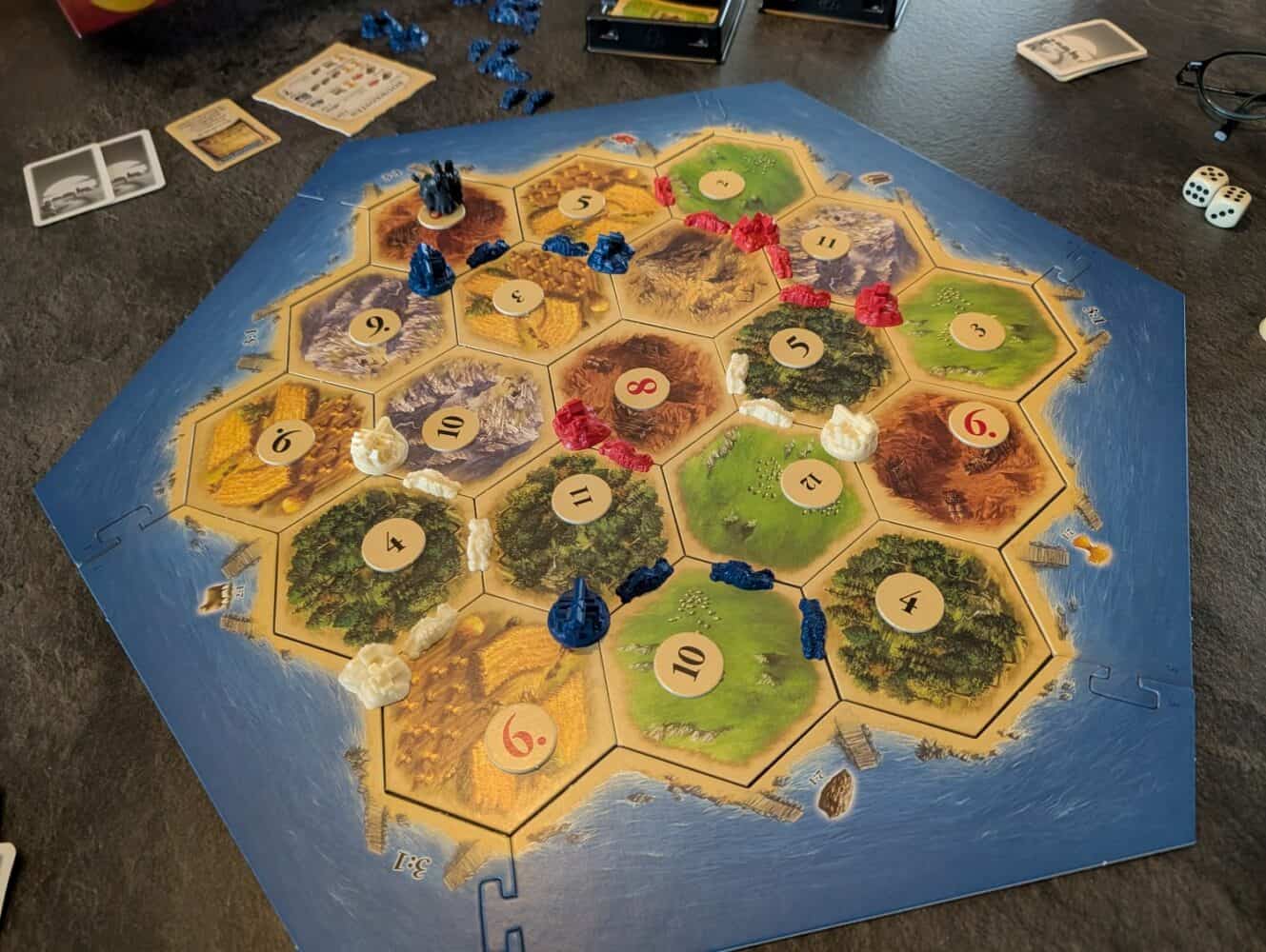 Een spel Catan in uitvoering, met het zeshoekige bord met nederzettingen en wegen in rood, blauw en wit, midden in het spel vastgelegd voor een recensie van de Google Pixel 10a, met grondstoffentegels, genummerde speelfiguren en speelkaarten op tafel.