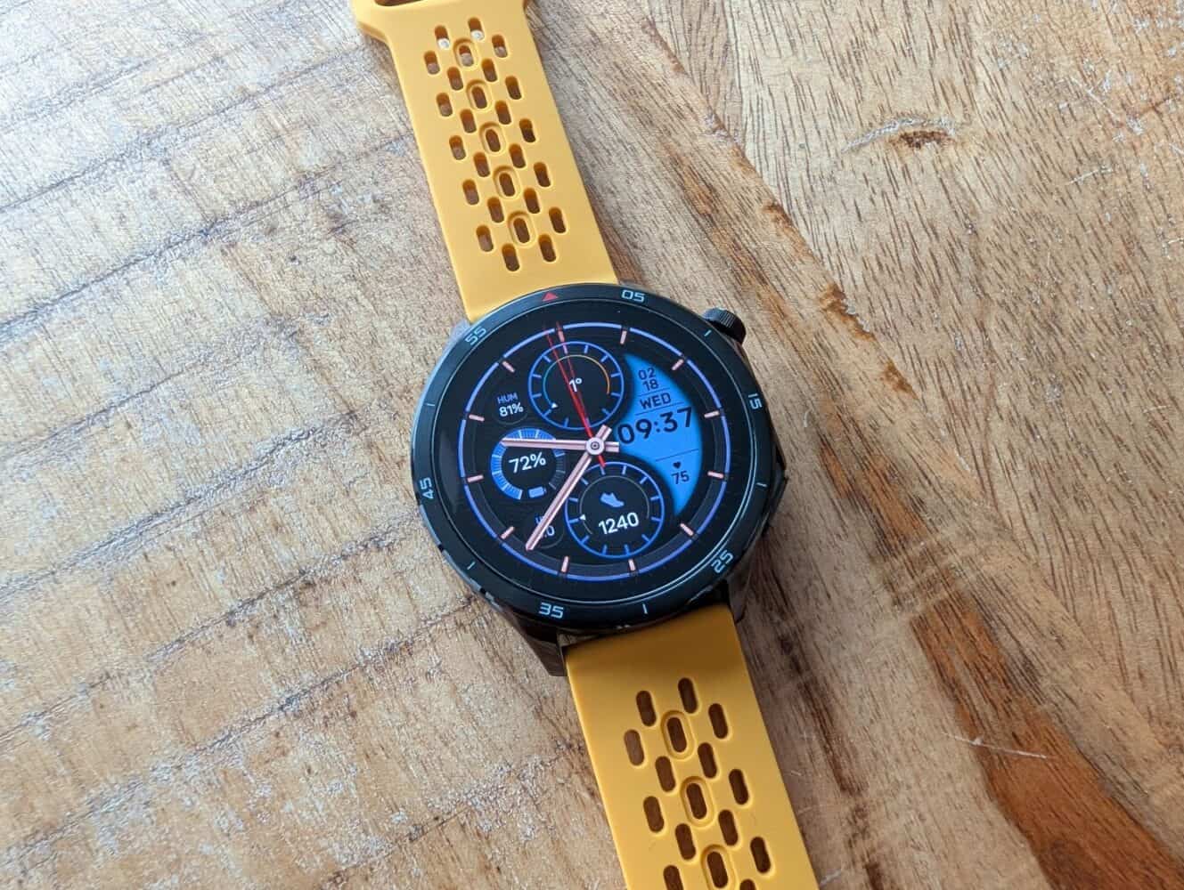 Een ronde smartwatch met een gele geperforeerde band toont de tijd, datum, batterijniveau, stappen en hartslag op een houten oppervlak - perfect om je conditie bij te houden terwijl je de laatste Google Pixel 10a review leest.