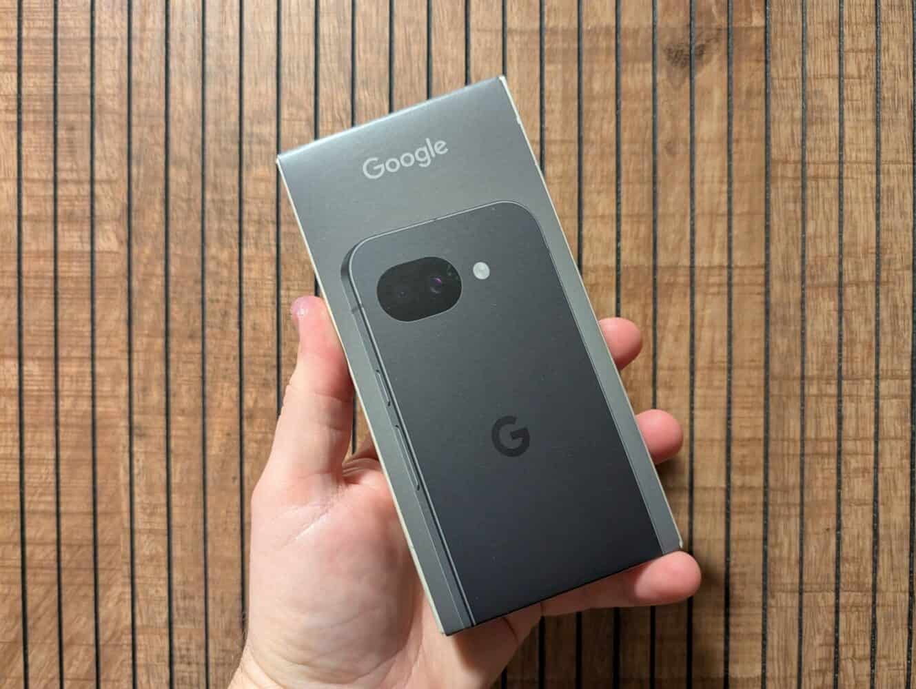 Een hand die een verzegelde doos van een Google smartphone vasthoudt op een houten oppervlak, klaar voor een uitgebreide Google Pixel 10a review.