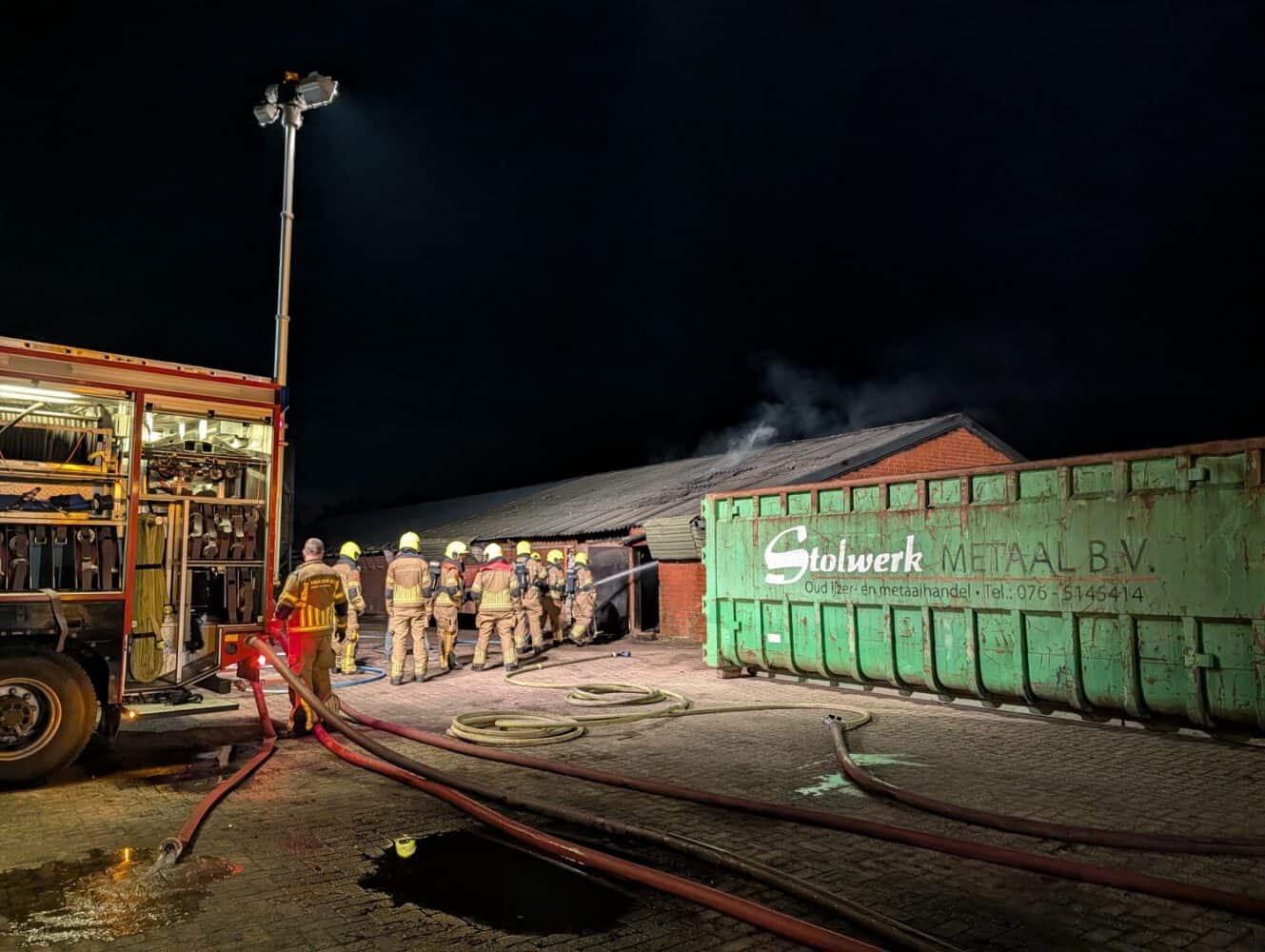 Brandweermannen in uitrusting staan bij een gebouw waar 's nachts rook vanaf het dak omhoog komt; een brandweerwagen en een grote groene metalen container staan vlakbij, haarscherp vastgelegd als een scène uit een Google Pixel 10a review.