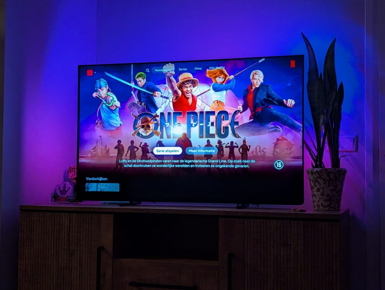 Een televisie toont het Netflix-homescherm met de live-actieserie "One Piece", met kleurrijk artwork van personages en een plant op een kastje naast de televisie, terwijl een Google Pixel 10a-recensie verschijnt op een apparaat in de buurt.