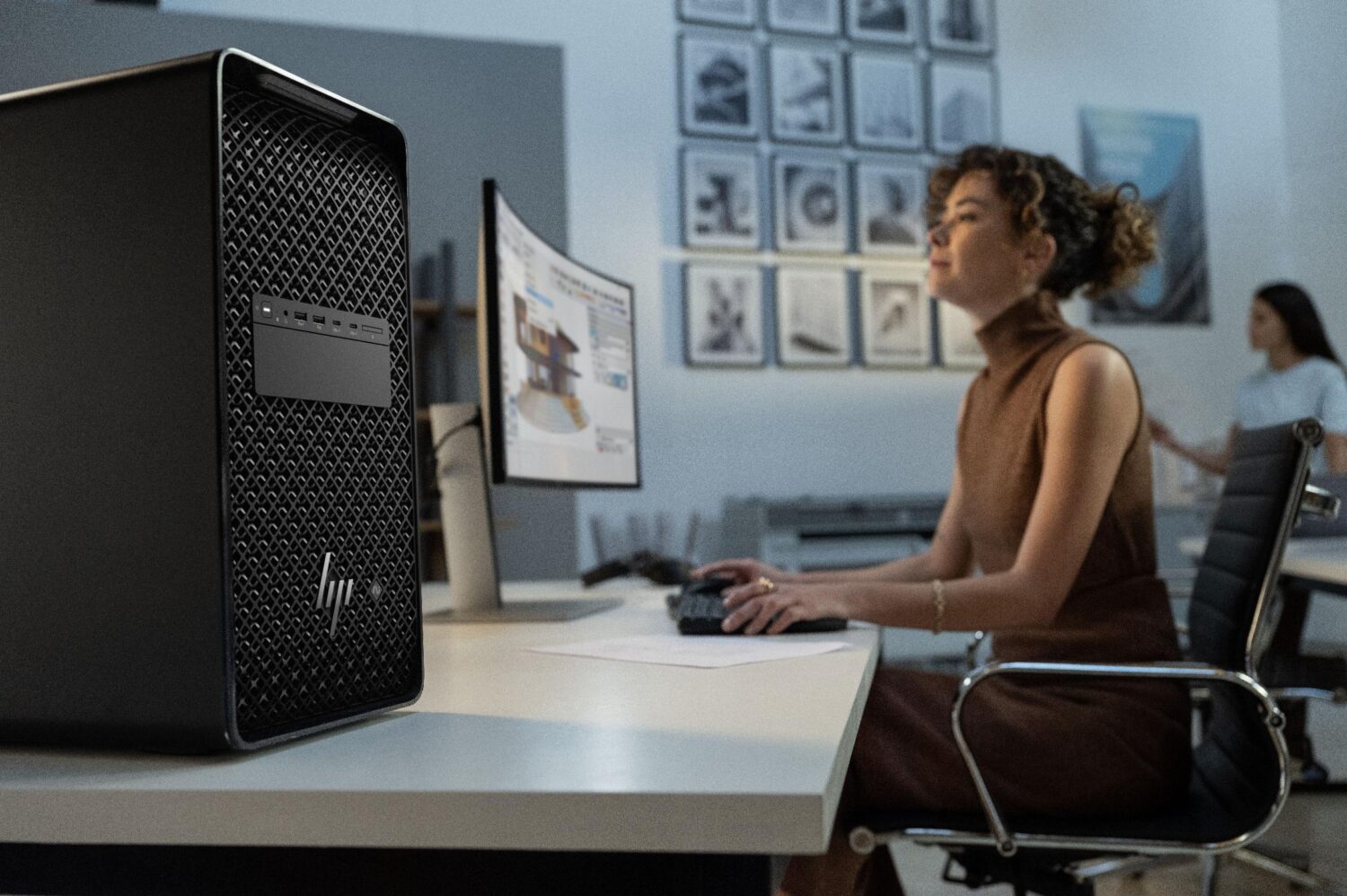 Een vrouw zit aan een bureau en gebruikt een computer met een groot bureaublad op de voorgrond; een andere persoon staat op de achtergrond.