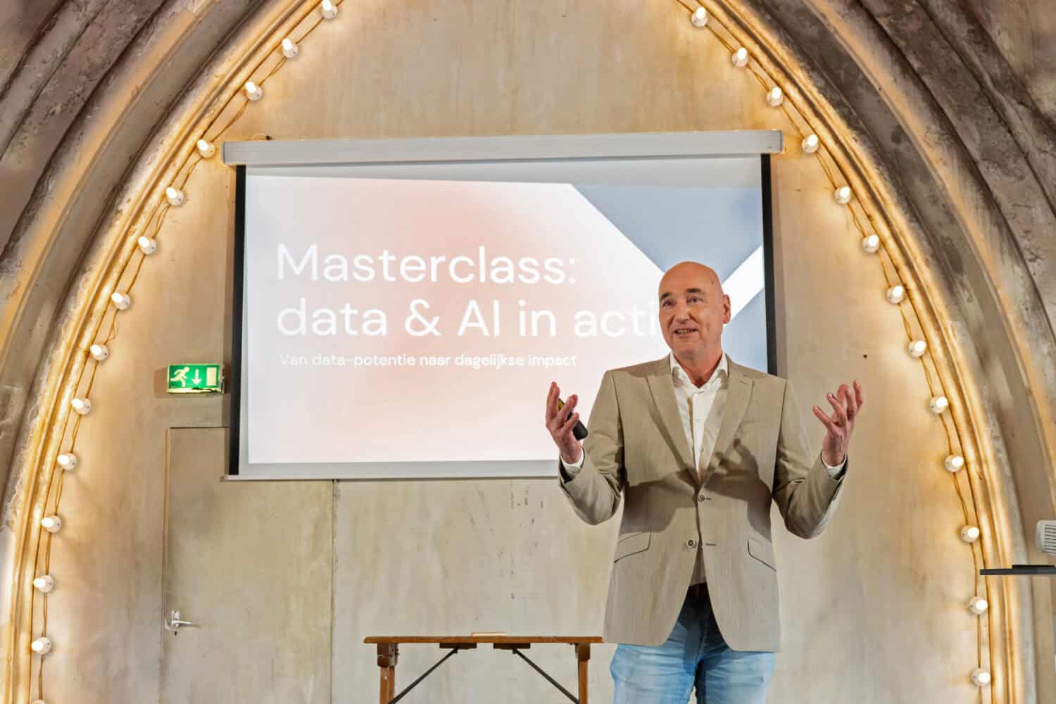 Een man in een beige blazer staat voor een scherm en presenteert een "Masterclass: data & AI in actie", waarin wordt besproken waarom BI en AI trajecten mislukken, in een kamer met gewelfde muren en strijklichtjes.