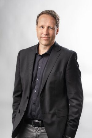 Man met lichtbruin haar draagt een zwart colbert, donker overhemd en grijze broek, staande tegen een effen witte achtergrond.