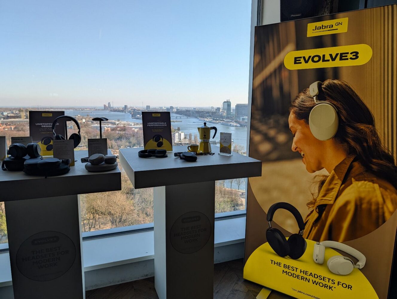 Display van Jabra Evolve hoofdtelefoons op tafels bij een raam, met uitzicht op de stad op de achtergrond en een grote Jabra-poster met een vrouw die een hoofdtelefoon draagt.