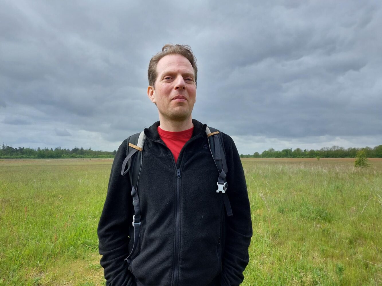 Een man met een zwarte jas en rugzak staat in een grasveld onder een bewolkte hemel, nadenkend over digitale soevereiniteit en de reis van open source migratie.