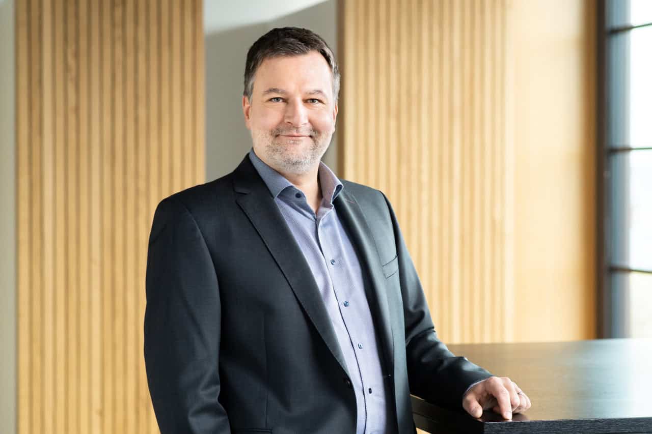 Een man in een donker pak en licht overhemd staat binnen naast een zwarte tafel en lacht naar de camera. Verticale houten panelen en grote ramen bevinden zich op de achtergrond.