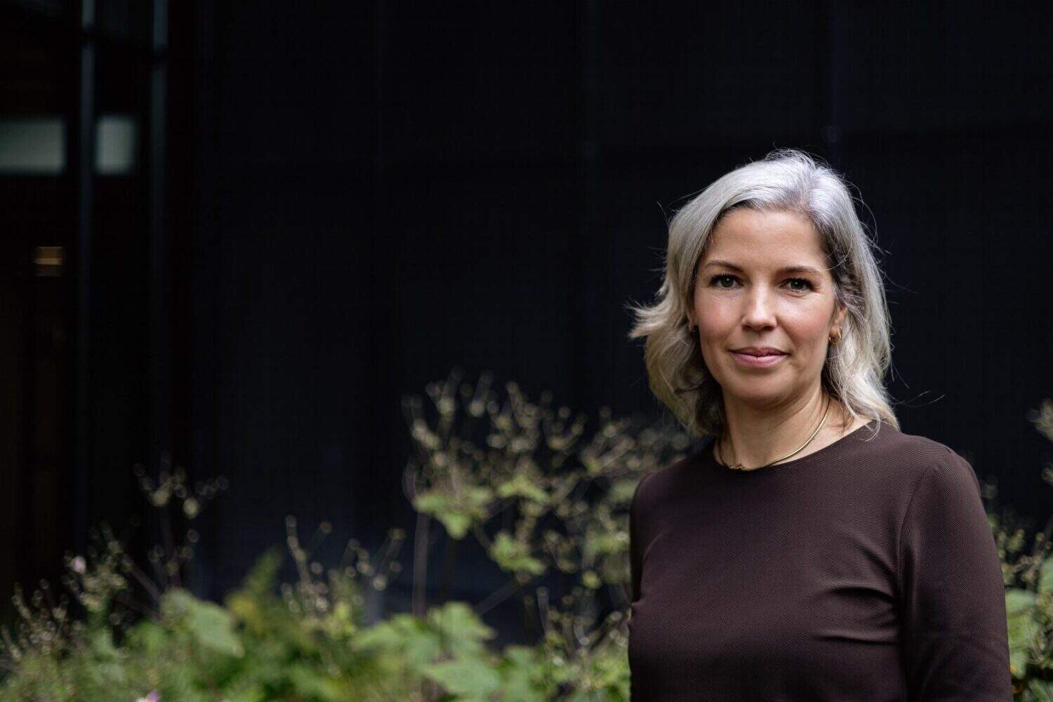 Een vrouw met grijs haar en een bruin topje staat buiten voor groen en een donker gebouw en belichaamt de ontspannen stijl van Northwave.