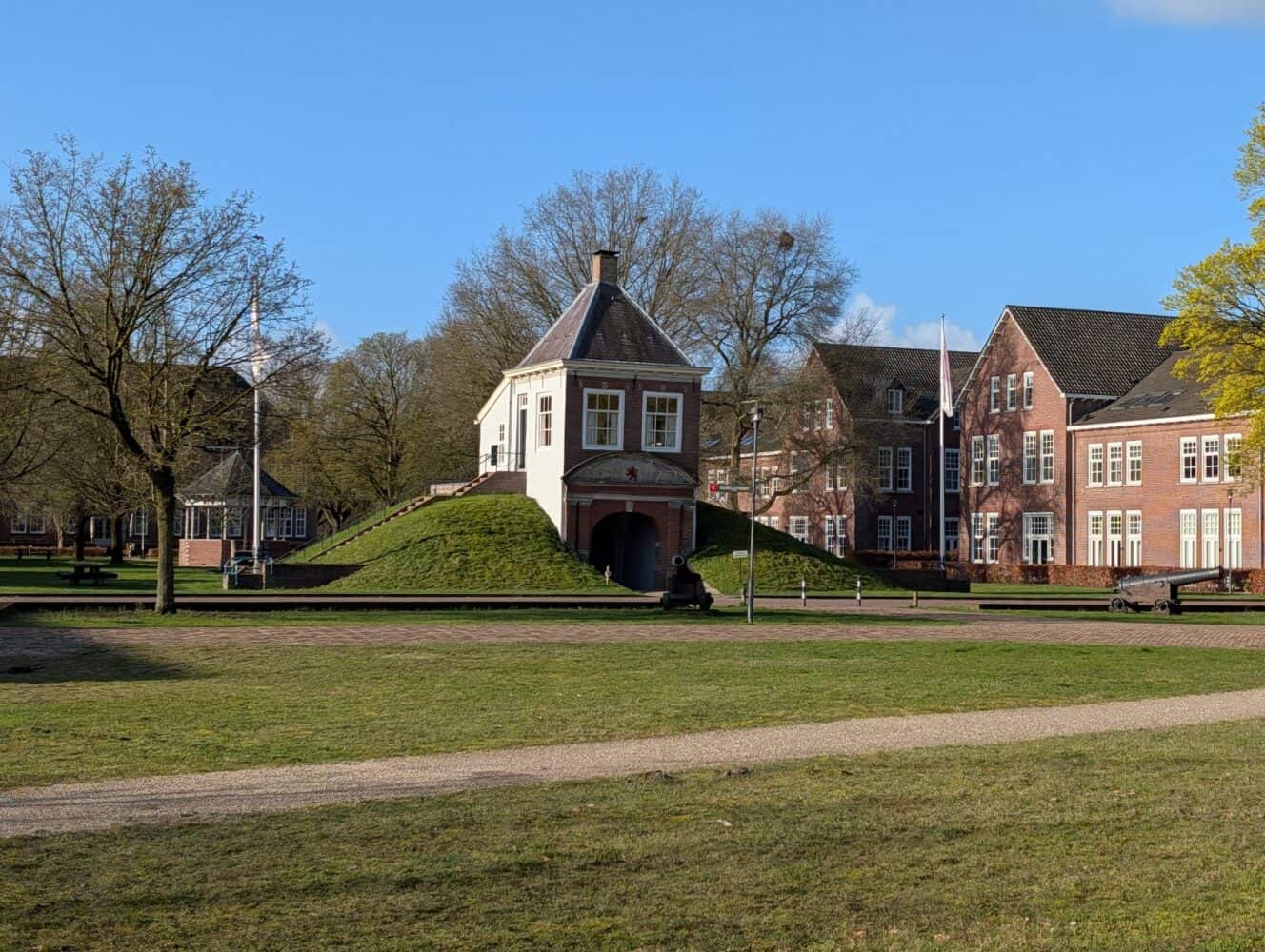 Een klein bakstenen gebouw met een steil dak staat op een grasheuvel, omringd door bomen en grenzend aan grotere bakstenen gebouwen onder een helderblauwe lucht, haarscherp vastgelegd zoals te zien in de Samsung Galaxy S26 Ultra review.