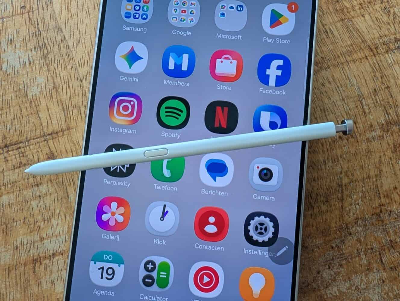 Een Samsung Galaxy S26 Ultra met verschillende app iconen ligt op een houten oppervlak, met een witte stylus pen diagonaal over het scherm - perfect voor een uitgebreide Samsung Galaxy S26 Ultra review.