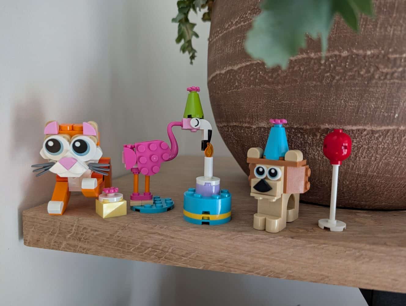 Vier kleine LEGO dierenfiguren - een kat, flamingo, vogel en een hond met een blauwe feestmuts - staan op een houten plank naast een rode lolly en een grote bloempot, waardoor een speels tafereel ontstaat dat perfect is om te fotograferen met je Samsung Galaxy S26 Ultra test sample.