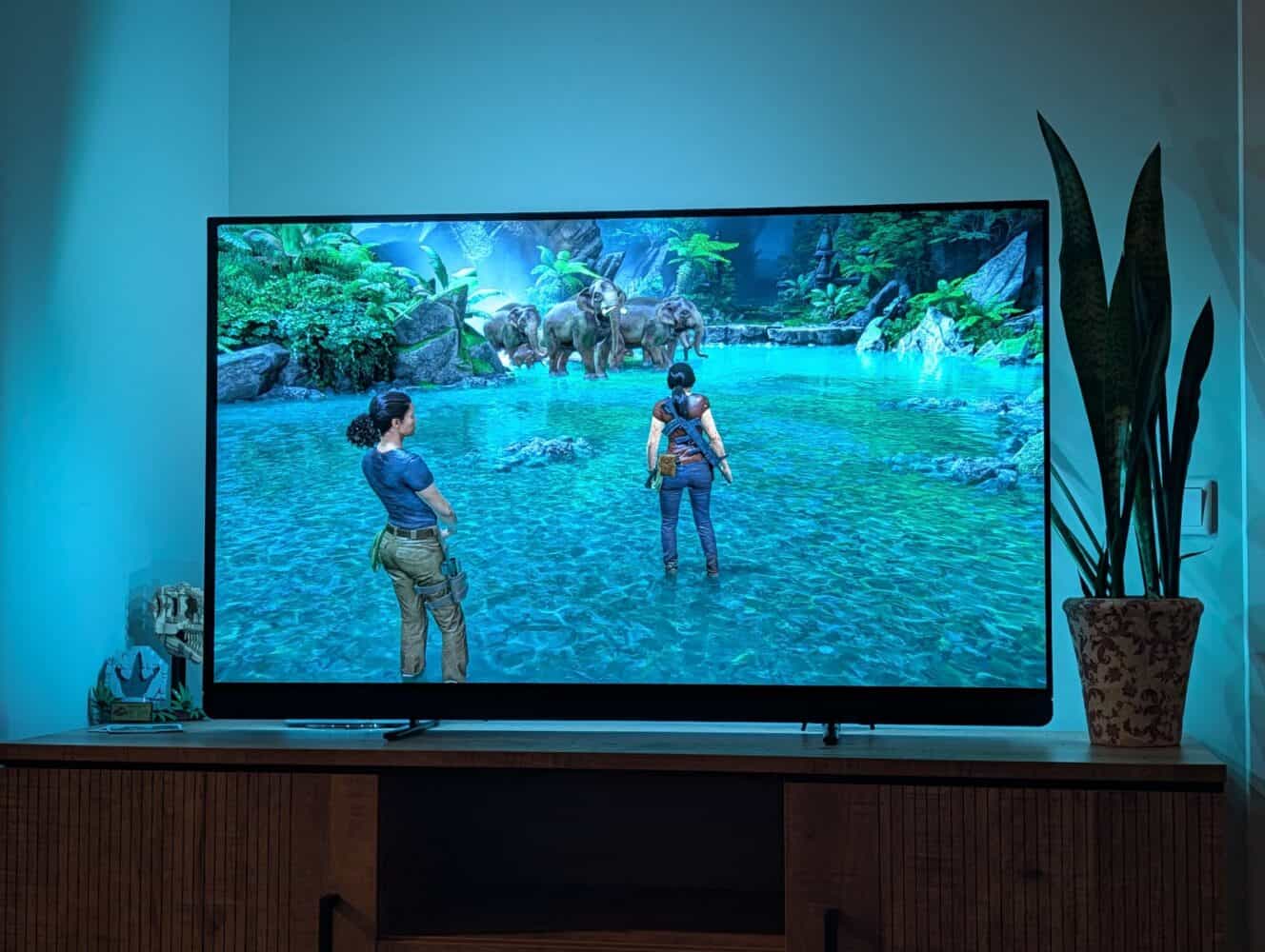 Een televisiescherm toont een scène van een videogame met twee personages die in ondiep water staan, tegenover een groep olifanten in een weelderige, tropische omgeving - perfect om de graphics te laten zien in een Samsung Galaxy S26 Ultra review.