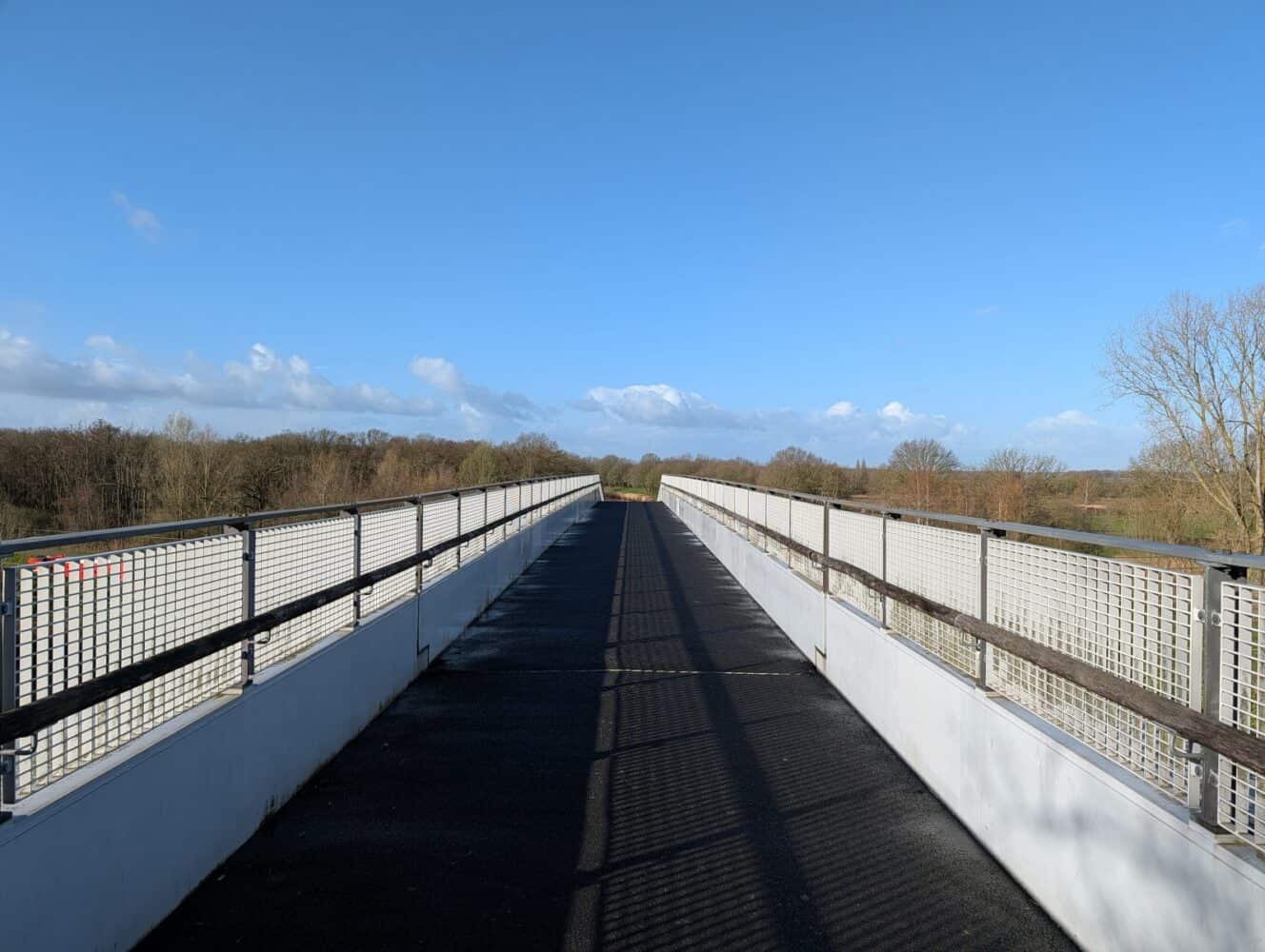 Een voetgangersbrug met metalen leuningen kruist een landschap van bomen en velden onder een helderblauwe lucht, vastgelegd in levendige details die doen denken aan foto's in een Samsung Galaxy S26 Ultra review.