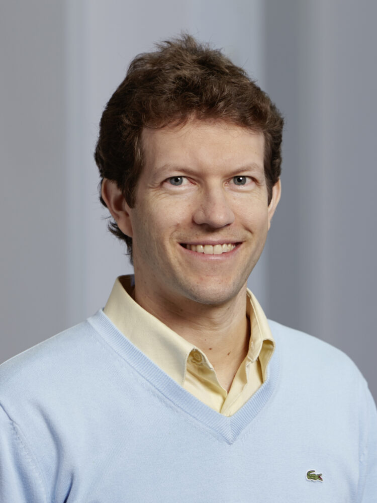 Een man met kort bruin haar, die een lichtblauwe trui draagt over een geel hemd met kraag, glimlacht naar de camera voor een effen achtergrond.