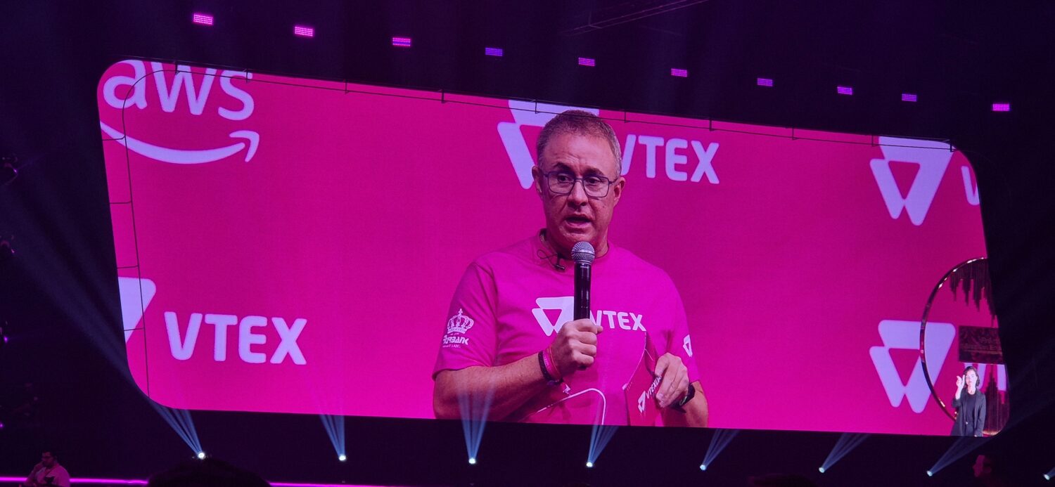 Een man in een roze VTEX-shirt spreekt op het podium met een microfoon en zijn beeld wordt geprojecteerd op een groot scherm achter hem met AWS- en VTEX-logo's, waarmee de impact van het VTEX enterprise commerce-platform in Europa wordt benadrukt.
