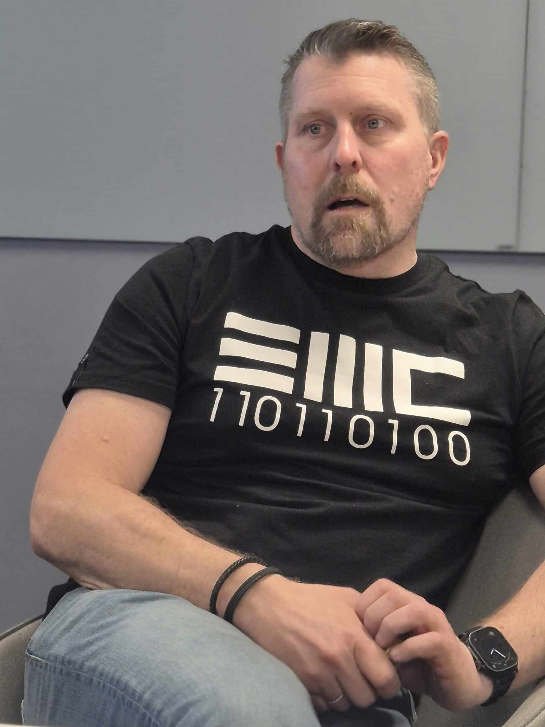 Een man met kort haar en een baard zit in een stoel, draagt een zwart T-shirt met een binaire code en een spijkerbroek, met zijn handen rustend in zijn schoot.