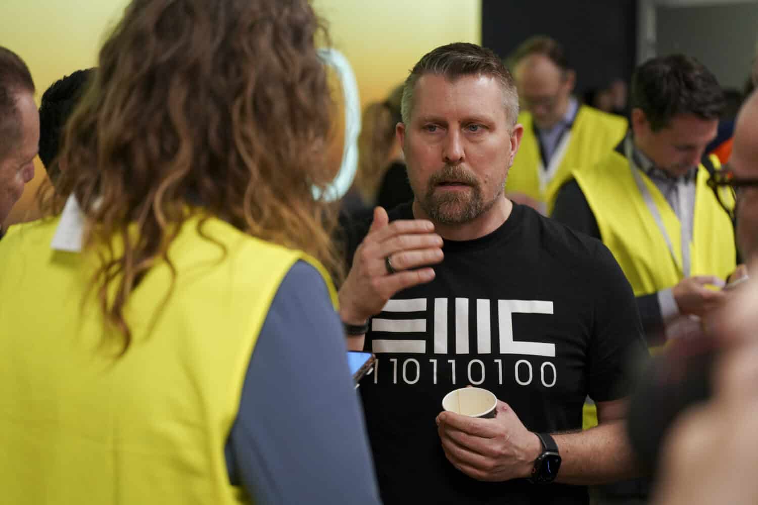 Een man in een zwart shirt met binaire cijfers praat met een persoon in een geel veiligheidsvest terwijl hij een papieren beker vasthoudt op een druk indoor evenement.