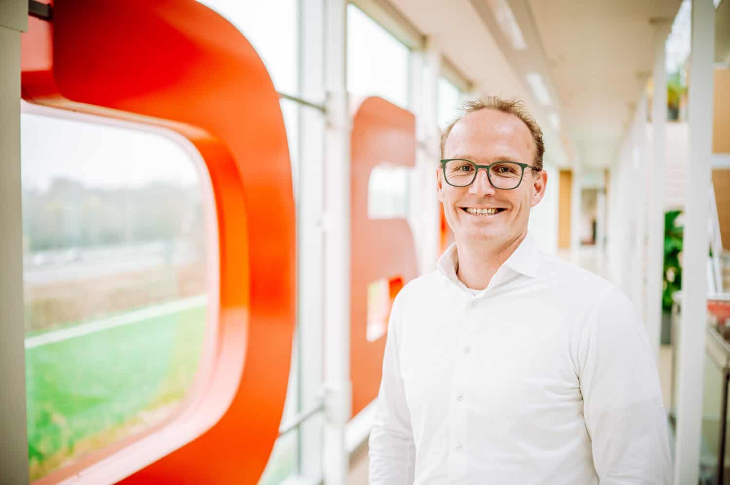 Een man met een bril en een wit overhemd staat glimlachend in een lichte gang met grote rode letters op het raam achter hem.