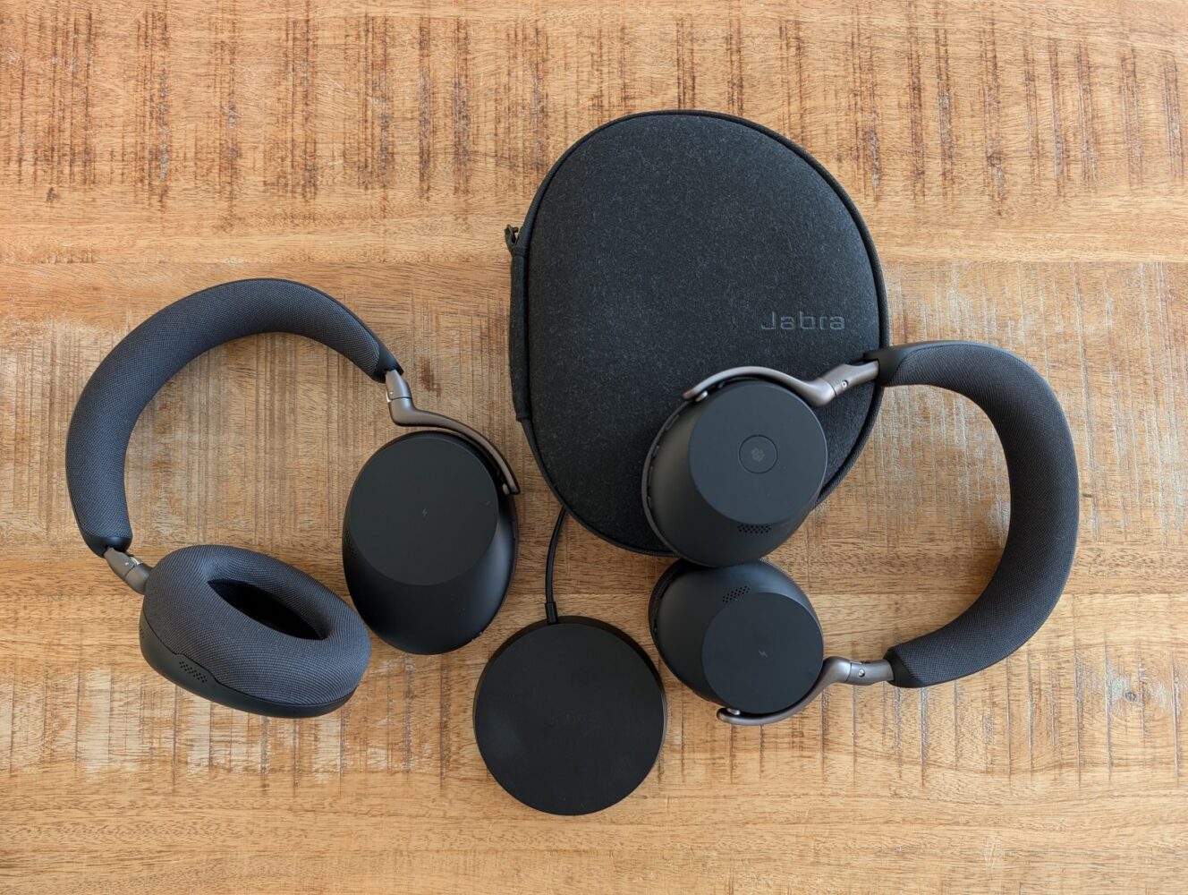 Twee paar zwarte over-ear hoofdtelefoons, een rond oplaadpad en een zwarte Jabra draagtas liggen op een houten oppervlak, perfect om uw eigen Jabra Evolve3 review op te zetten.