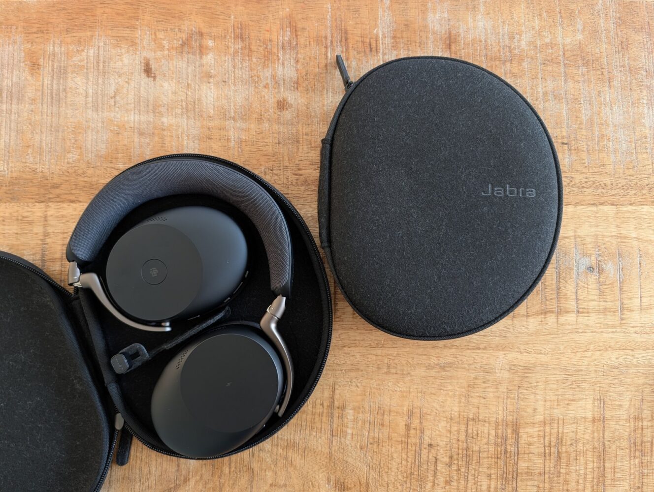 Een paar zwarte Jabra headphones in een open etui ligt naast een gesloten Jabra etui op een houten oppervlak, zoals te zien is in onze Jabra Evolve3 review.