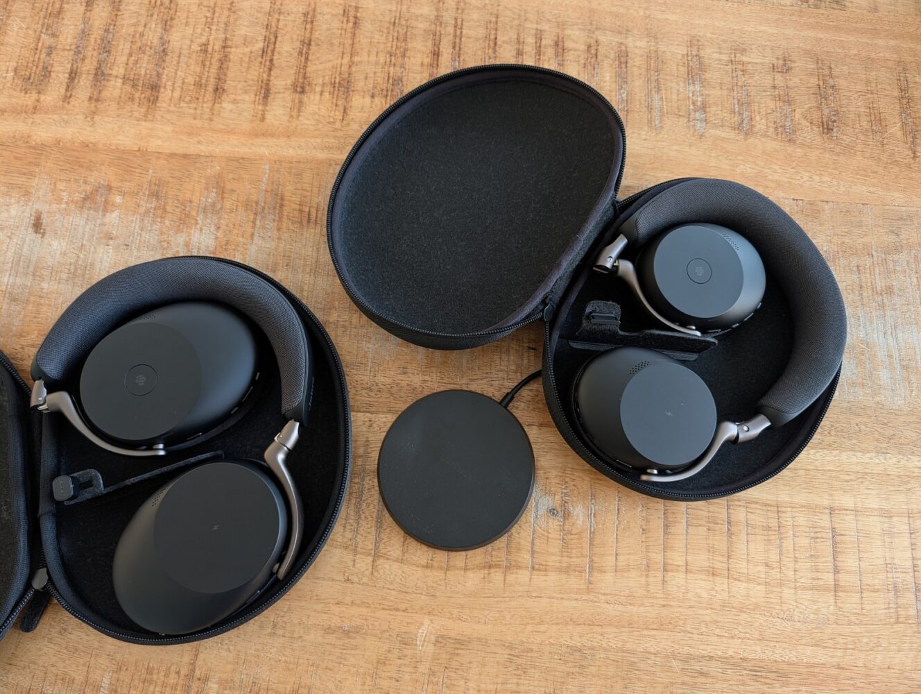 Twee paar zwarte over-ear hoofdtelefoons in open hoesjes en een rond zwart oplaadkussen zijn geplaatst op een houten oppervlak, wat het slanke ontwerp benadrukt dat te zien is op veel foto's van Jabra Evolve3 reviews.