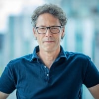 Een man met krullend haar en een bril in een donkerblauwe polo staat voor een wazige achtergrond.