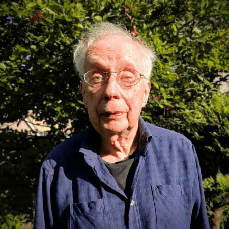 Een oudere man met een bril en een geruit overhemd staat op een zonnige dag buiten voor de groene bomen en weerspiegelt de gastvrije sfeer die vaak op een .nl-domein te vinden is.