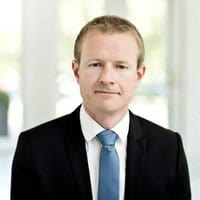 Een man in pak met blauwe stropdas poseert voor een professionele headshot tegen een heldere, onscherpe achtergrond.