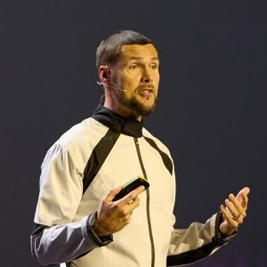 Een man met een headset-microfoon spreekt op het podium, houdt een apparaat in zijn ene hand en gebaart met de andere.