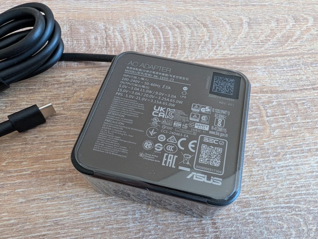 Een close-up van een ASUS USB-C laptop AC-adapter met technische specificaties en certificeringslogo's zichtbaar op het label, geplaatst op een houten oppervlak.