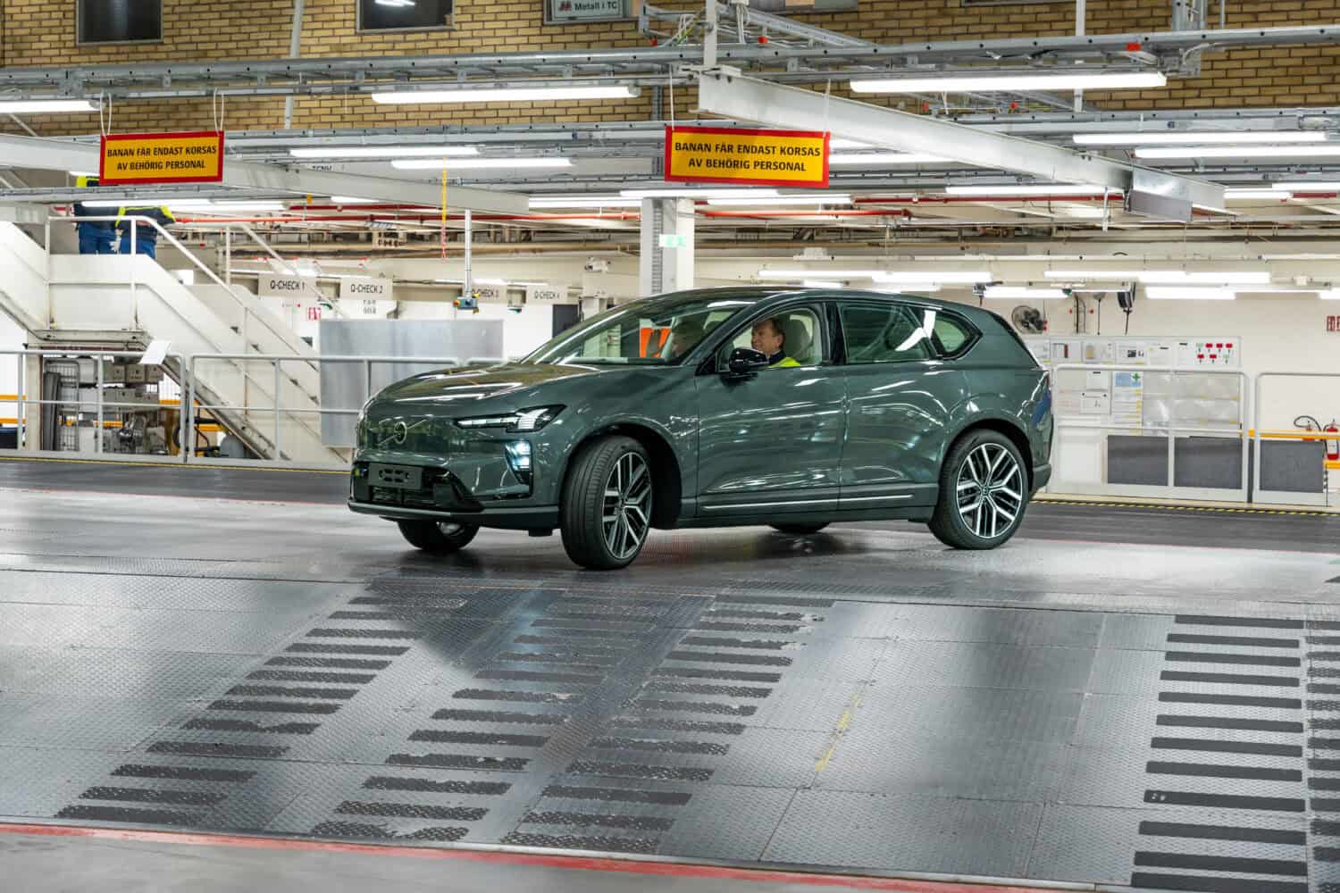 Een grijze elektrische SUV staat in een moderne autofabriek, omringd door industriële apparatuur en veiligheidsborden boven het hoofd.