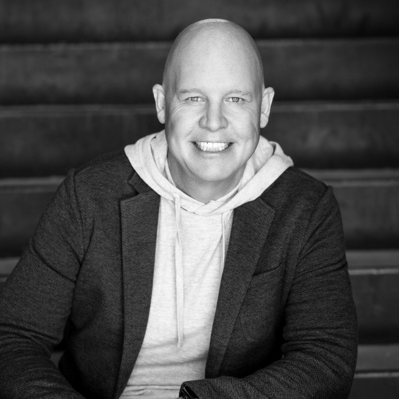 Een kale man, die de cyberbeveiligingswerkers vertegenwoordigt, draagt een capuchon en blazer terwijl hij op een trapje zit en naar de camera glimlacht op een zwart-witfoto.