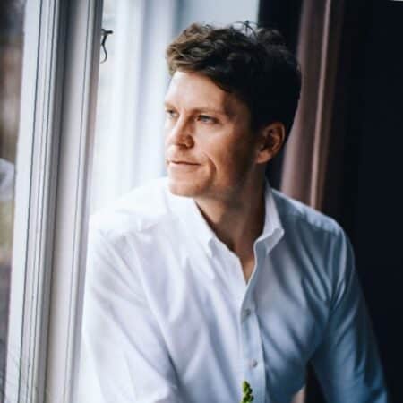 Een man in een wit overhemd kijkt met een bedachtzame uitdrukking uit een raam. Natuurlijk licht verlicht zijn gezicht.