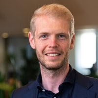 Een man met kort blond haar en een getrimde baard draagt een donker colbert en glimlacht naar de camera in een overdekte omgeving.