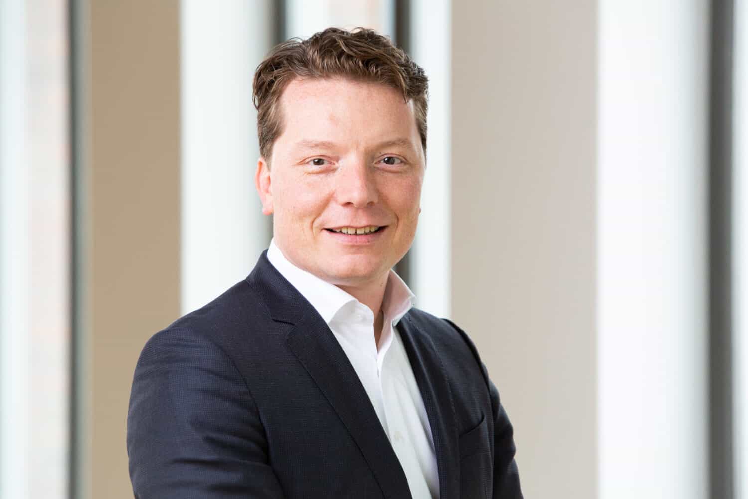 Een man in een donker pak en wit overhemd staat te glimlachen in een helder, modern kantoor met grote ramen op de achtergrond.