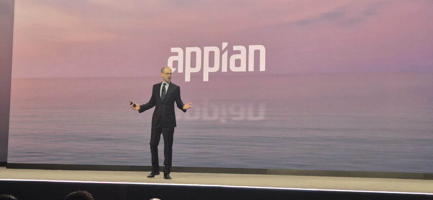 Een man in pak staat op het podium voor een groot scherm waarop "Appian" te zien is, met AI procesintegratie tegen een serene waterachtergrond om bedrijfswaarde te benadrukken.