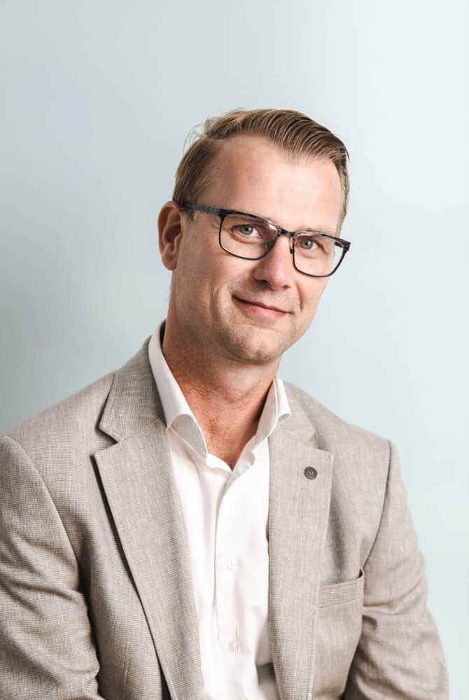 Een man met een bril en een lichtgrijs colbert over een wit overhemd zit tegen een lichtblauwe achtergrond, kijkt naar de camera en glimlacht lichtjes.