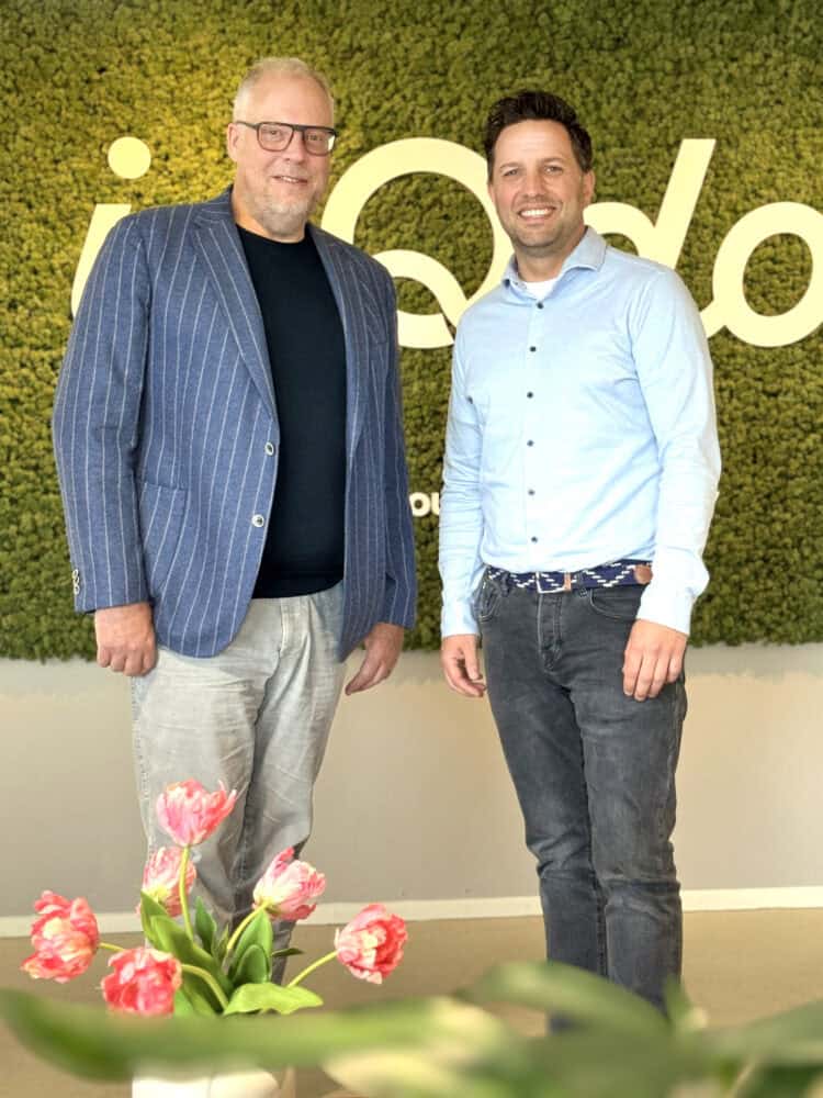 Twee mannen staan en glimlachen binnenshuis voor een groene mosmuur met witte tekst, met een vaas roze tulpen op de voorgrond.