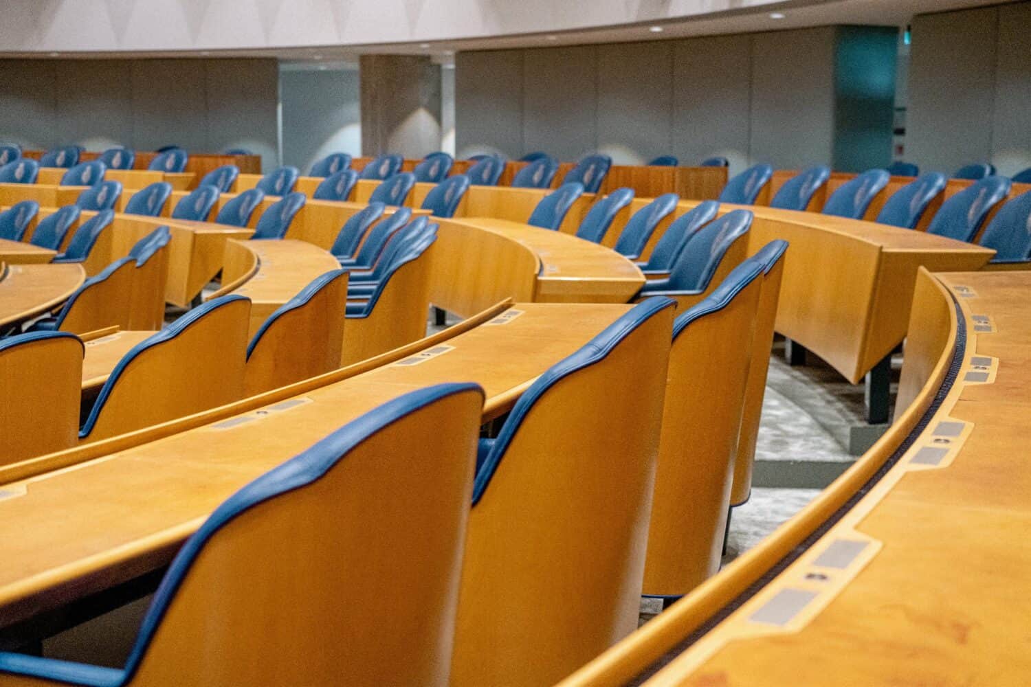 Rijen lege houten stoelen met blauwe kussens zijn gerangschikt in een gebogen patroon in een moderne collegezaal, waardoor de perfecte omgeving ontstaat voor discussies of Kamervragensessies.
