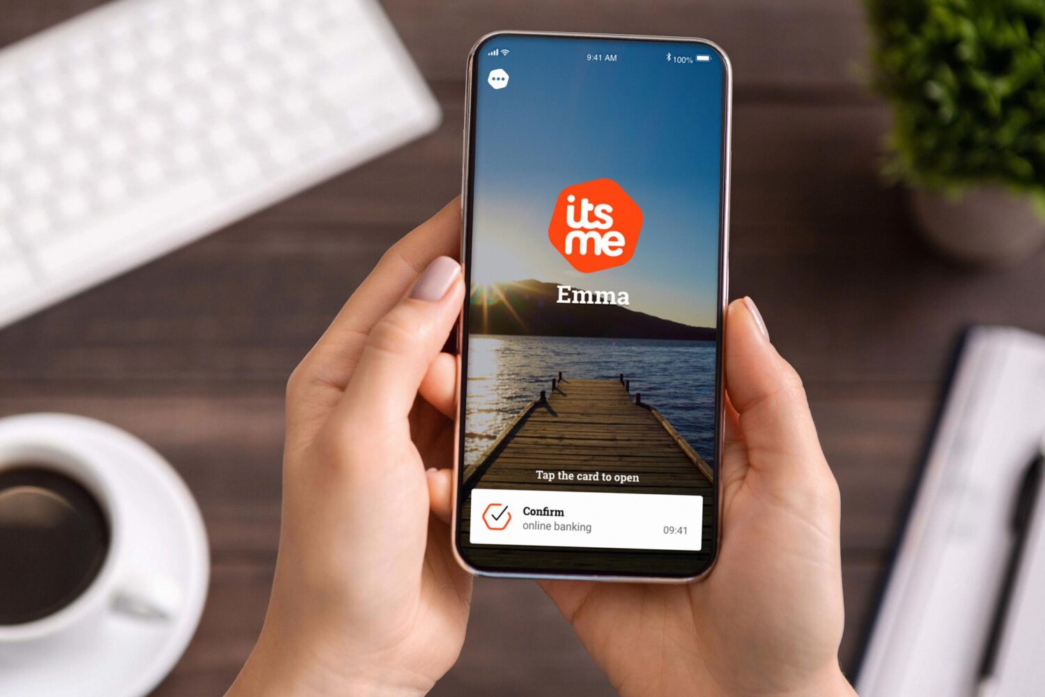De persoon houdt een smartphone vast met daarop de app "itsme" met een foto op het dock, de naam "Emma" en een vraag om online bankieren te bevestigen onderaan het scherm.