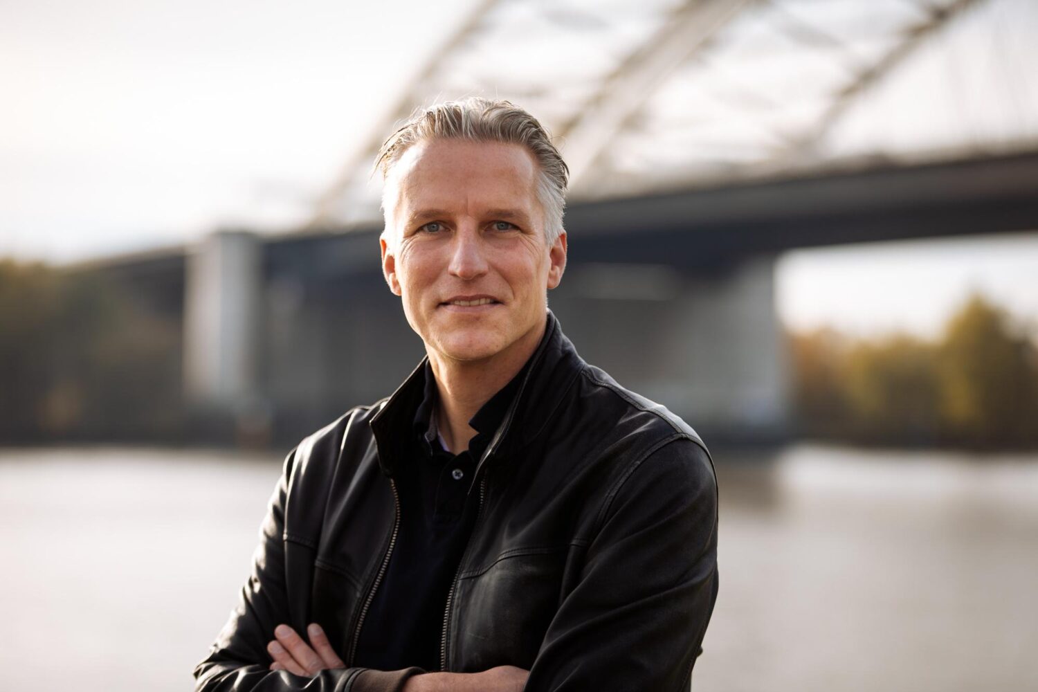 Een man met kort grijs haar, gekleed in een zwart leren jack, staat buiten met zijn armen over elkaar, met op de achtergrond een rivier en de indrukwekkende Office.eu-brug.