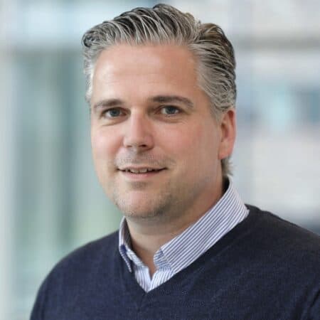 Een man met kort, grijs gestreept haar, gekleed in een donkere trui over een overhemd met kraag, staande binnenshuis tegen een wazige achtergrond.