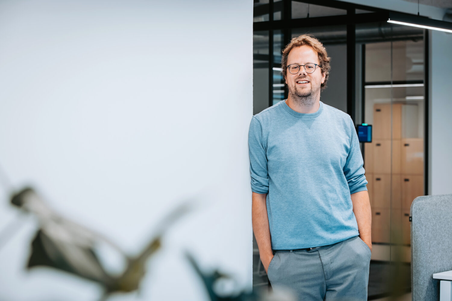 Een man met een bril en een blauwe trui staat nonchalant met zijn handen in zijn zakken in een modern kantoor, glimlacht en kijkt een beetje opzij.